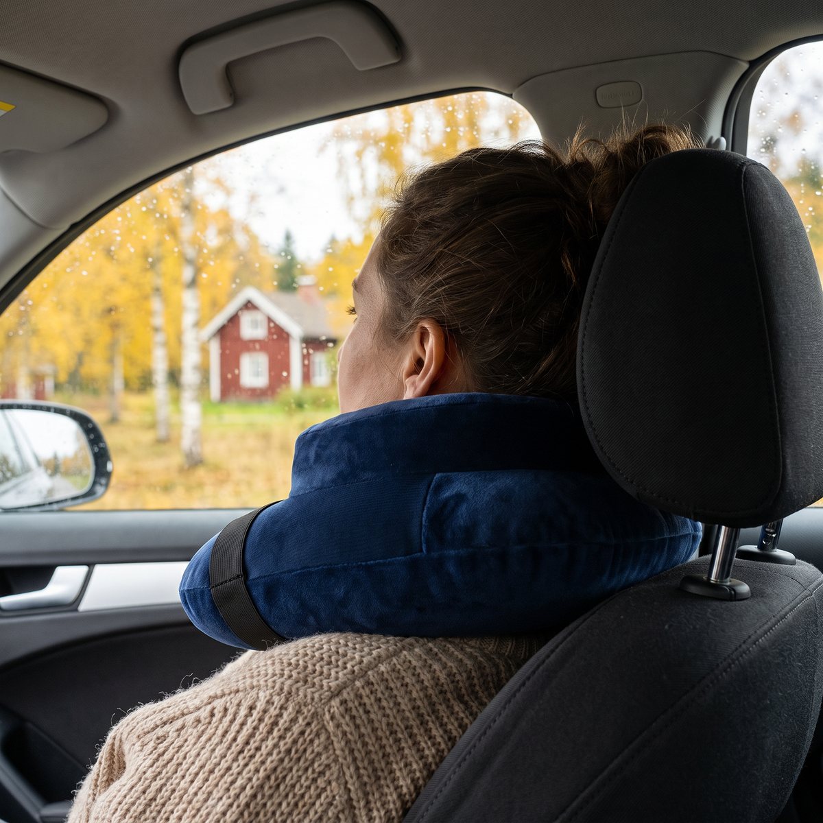 Marinblå uppblåsbar nackkudde med svart rem på en passagerare i framsätet, med gula björkar och en röd stuga utanför bilrutan