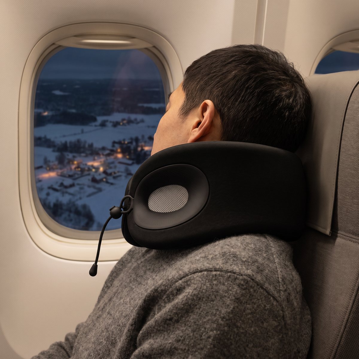 Svart nackkudde med gråa ventilpaneler och snöre under hakan på en sovande flygresenär vid fönsterplatsen, med snötäckt svenskt landskap i kvällsljus nedanför