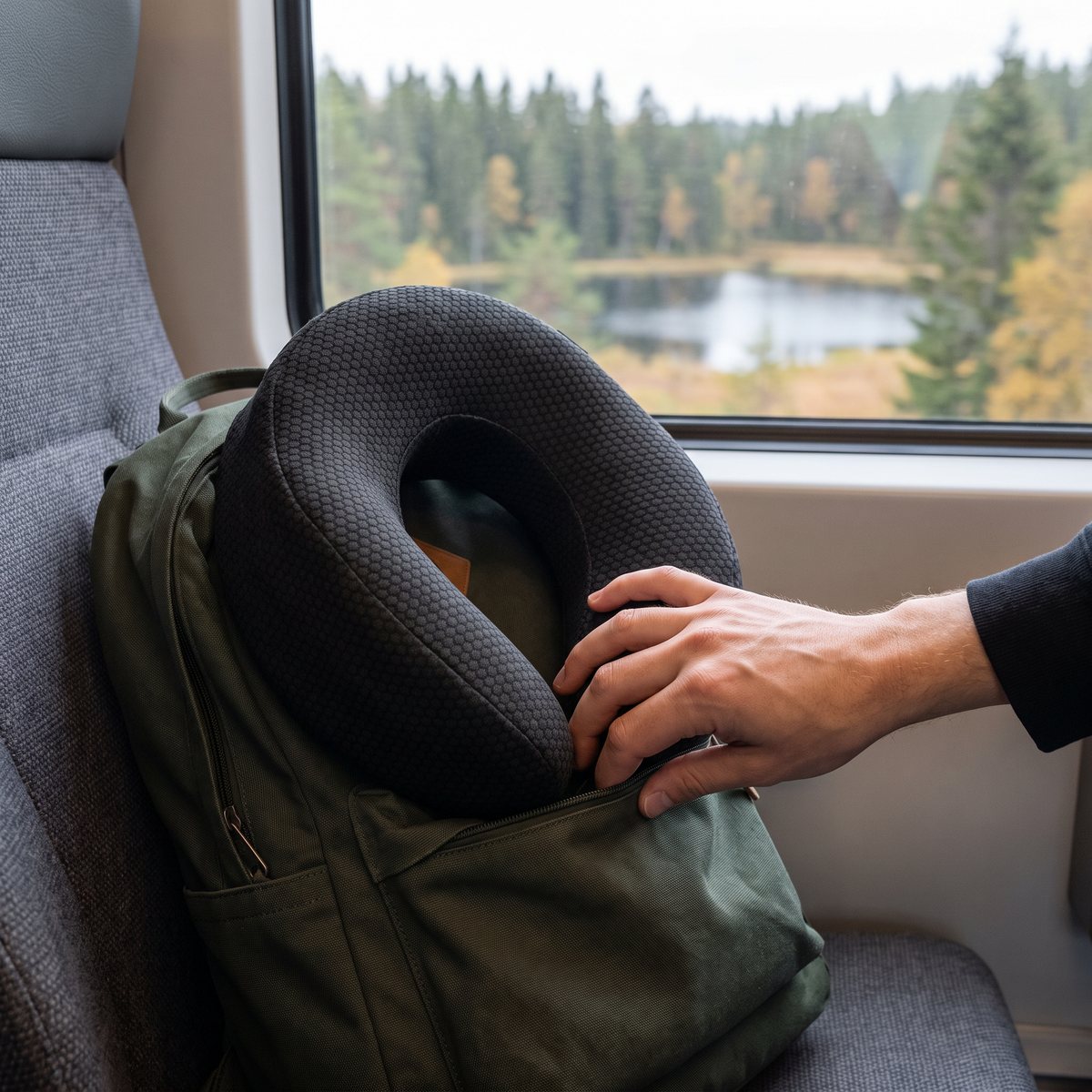 Svart nackkudde med bikakemönster som stoppas ner i en grön ryggsäck på ett tågsäte, med skog och sjö utanför fönstret