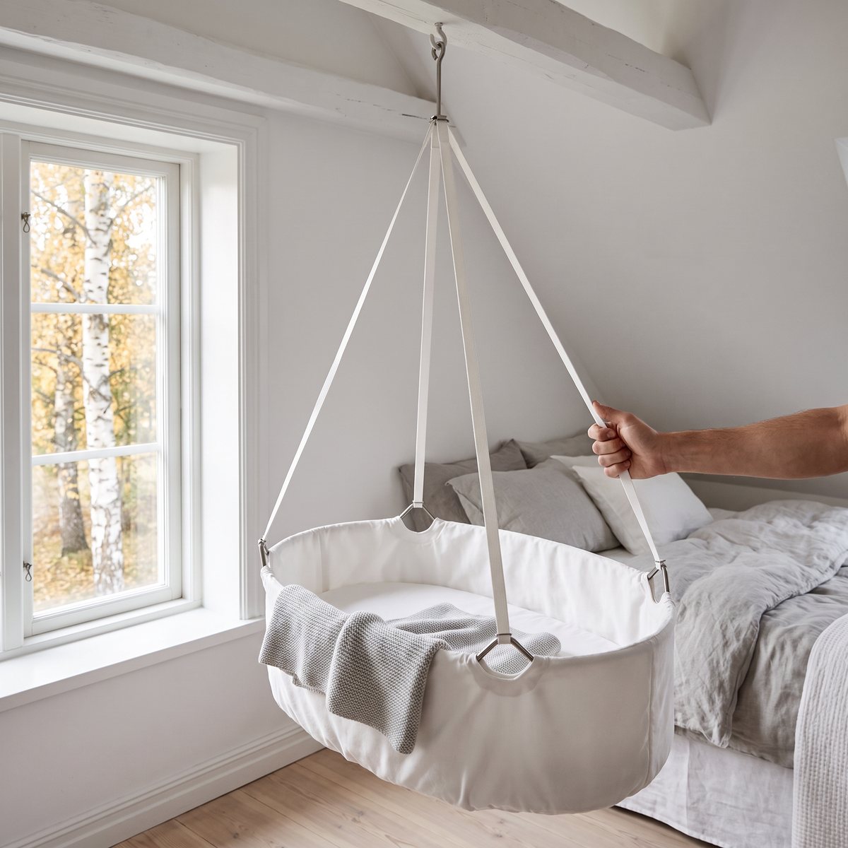 Vit Leander Classic hängvagga i ekologisk bomull som hänger från en takkrok i ett ljust svenskt sovrum, en hand ger den en lätt knuff