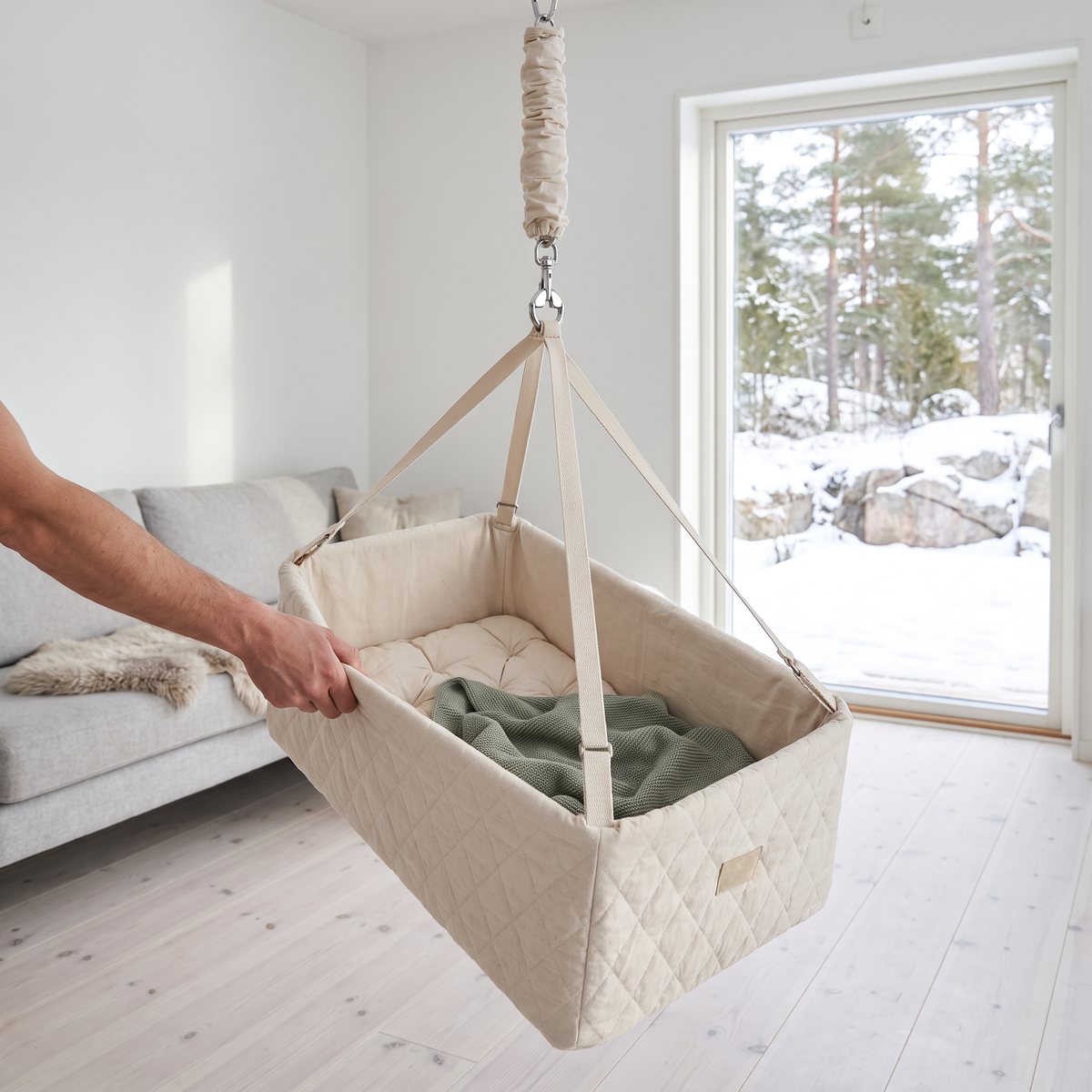 Krämvit quiltad Moonboon Vagga Nature på fjäder i ett ljust svenskt vardagsrum, en hand gungar vaggan för hand