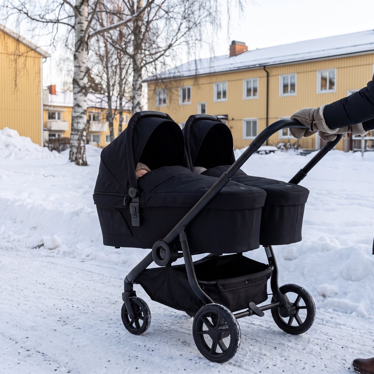 Beemoo Pro Twin 2 med två svarta liggdelar skjuts på en snöröjd gångväg vid gula flerfamiljshus en vinterdag