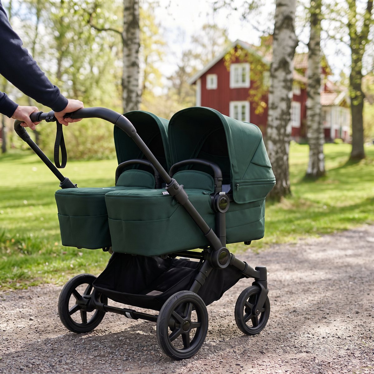 Bugaboo Donkey 6 i grönt med två liggdelar sida vid sida skjuts på en grusgång i en svensk park med björkar och ett rött trähus