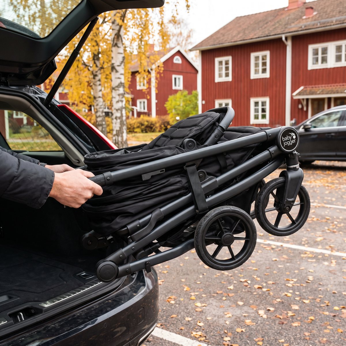 Hopfälld svart Baby Jogger City Mini GT2 Double lyfts in i bagageutrymmet på en bil framför röda trähus