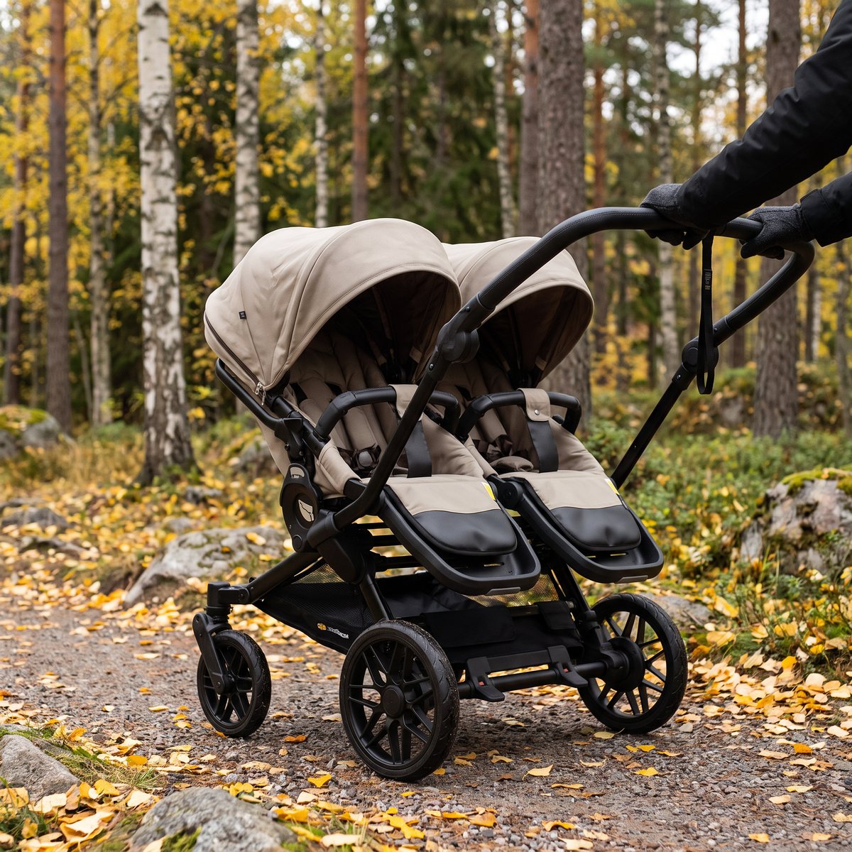 Beige Emmaljunga Double Sento sittvagn för två barn skjuts på en grusstig i en svensk höstskog med björk och tall