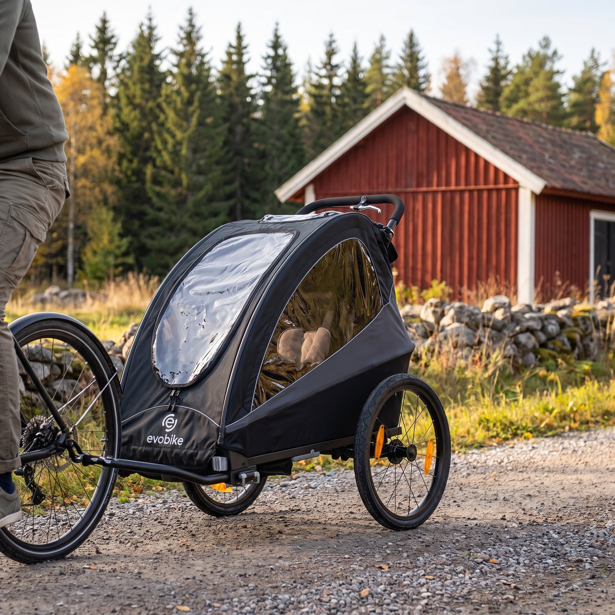 Svartgrå Evobike Yxlan med stora luftdäck dragen efter en cykel på en svensk grusväg vid en röd lada och stenmur.