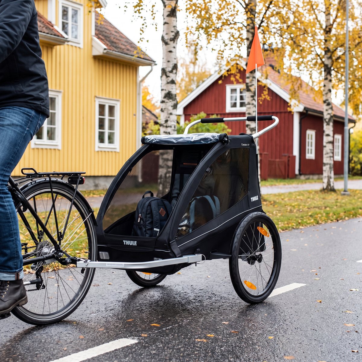Svart Thule Coaster XT med cykelflagga dragen efter en cykel på en cykelbana mellan gula och röda trähus en höstdag.