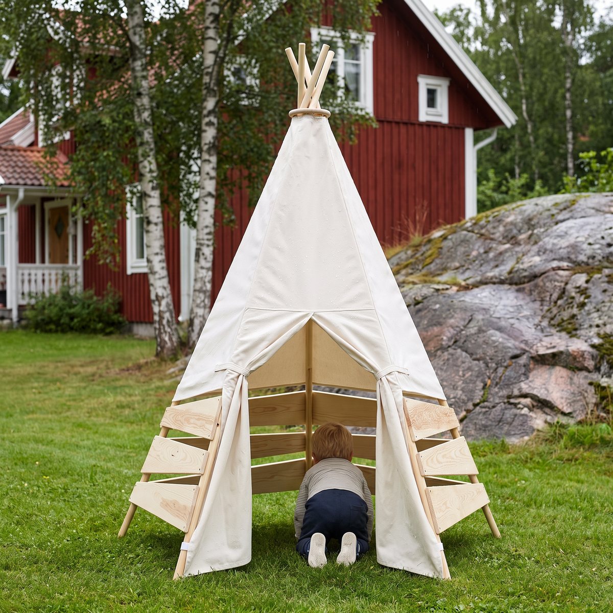 Vitt tipitält med trästomme och träribbor på gräsmattan framför ett rött svenskt trähus med björkar, ett barn kryper in genom öppningen