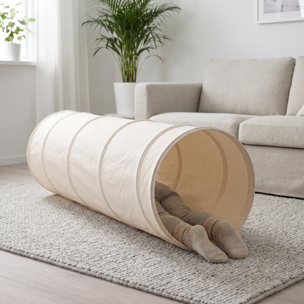 Naturvit lektunnel i bomull på en grå ullmatta i ett ljust svenskt vardagsrum, med ett barns ben i stickade sockor i öppningen