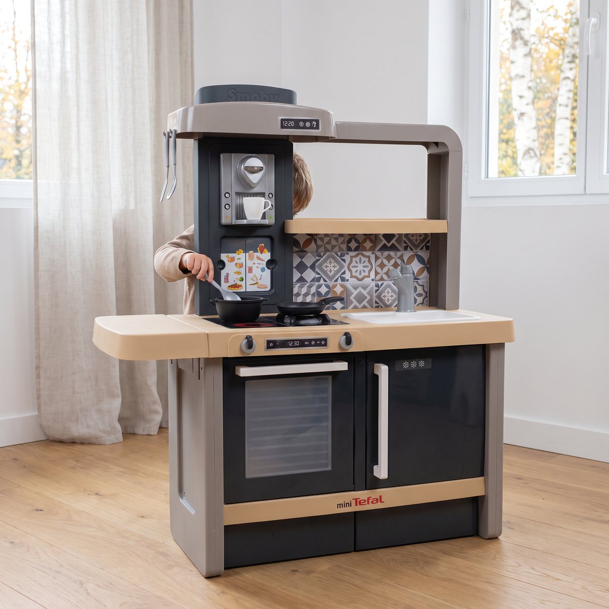 Smoby Tefal Evolutive leksakskök i grått och sandfärgat med kaffemaskin, spis, ugn och kylskåp i ett ljust svenskt barnrum med trägolv och björkar utanför fönstret