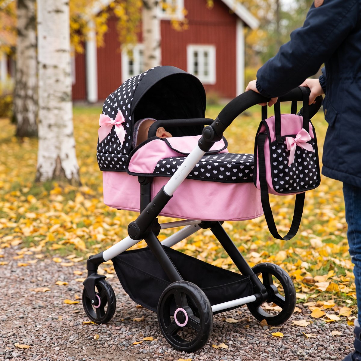 Bayer Design Xeo dockvagn i svart och rosa med skötväska på handtaget, på en grusgång bland björkar och höstlöv framför ett rött trähus