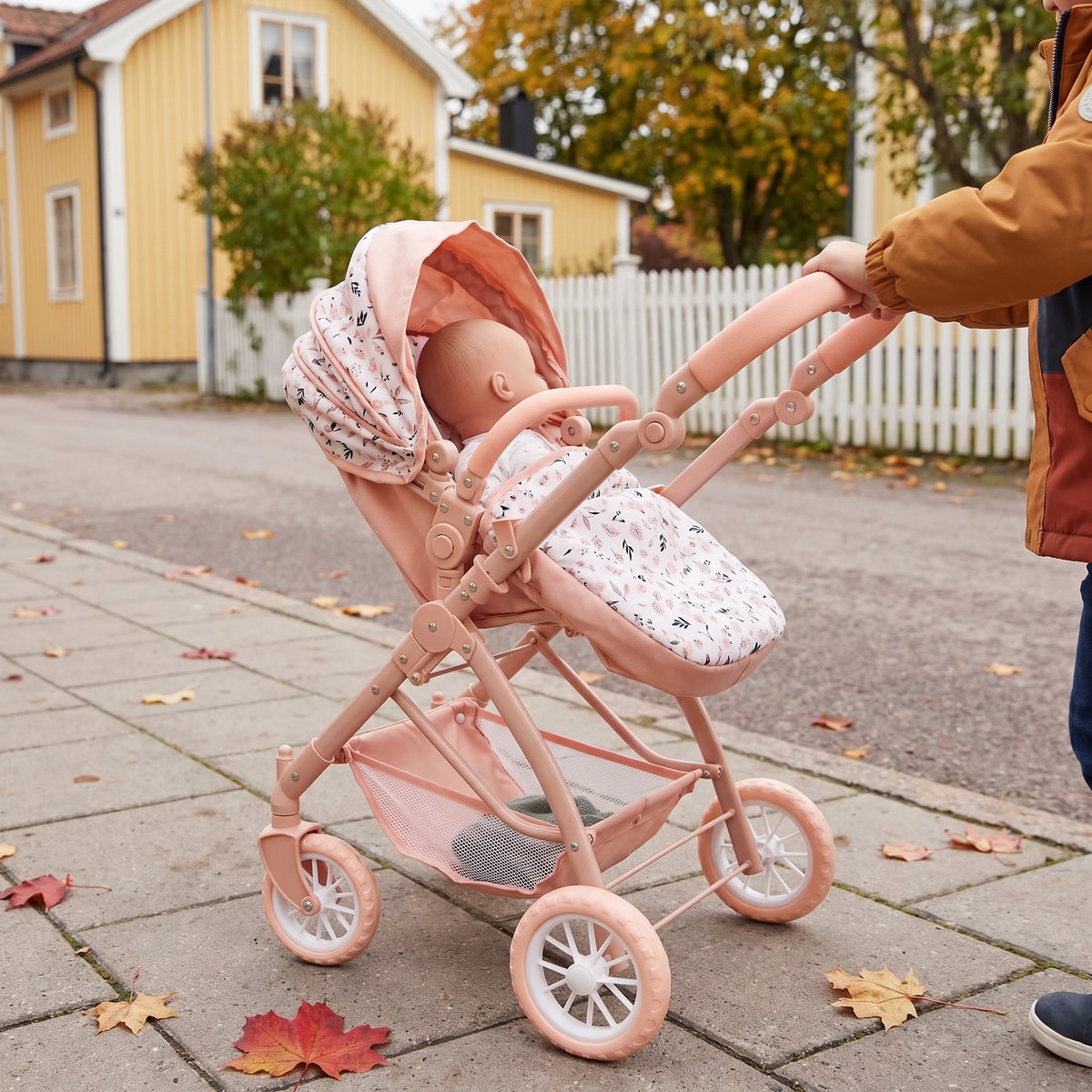 Cloudberry Castle Konvertibel Kanin i persikorosa med sitsen vänd mot barnet som skjuter vagnen på en trottoar bland gula trähus på hösten