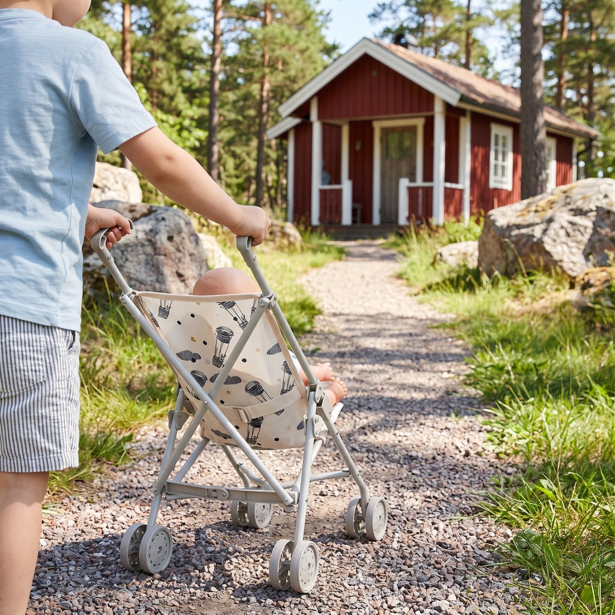 Skrållan docksulky med ballongmönster skjuts av ett barn på en grusstig mot en röd stuga bland tallar och granithällar