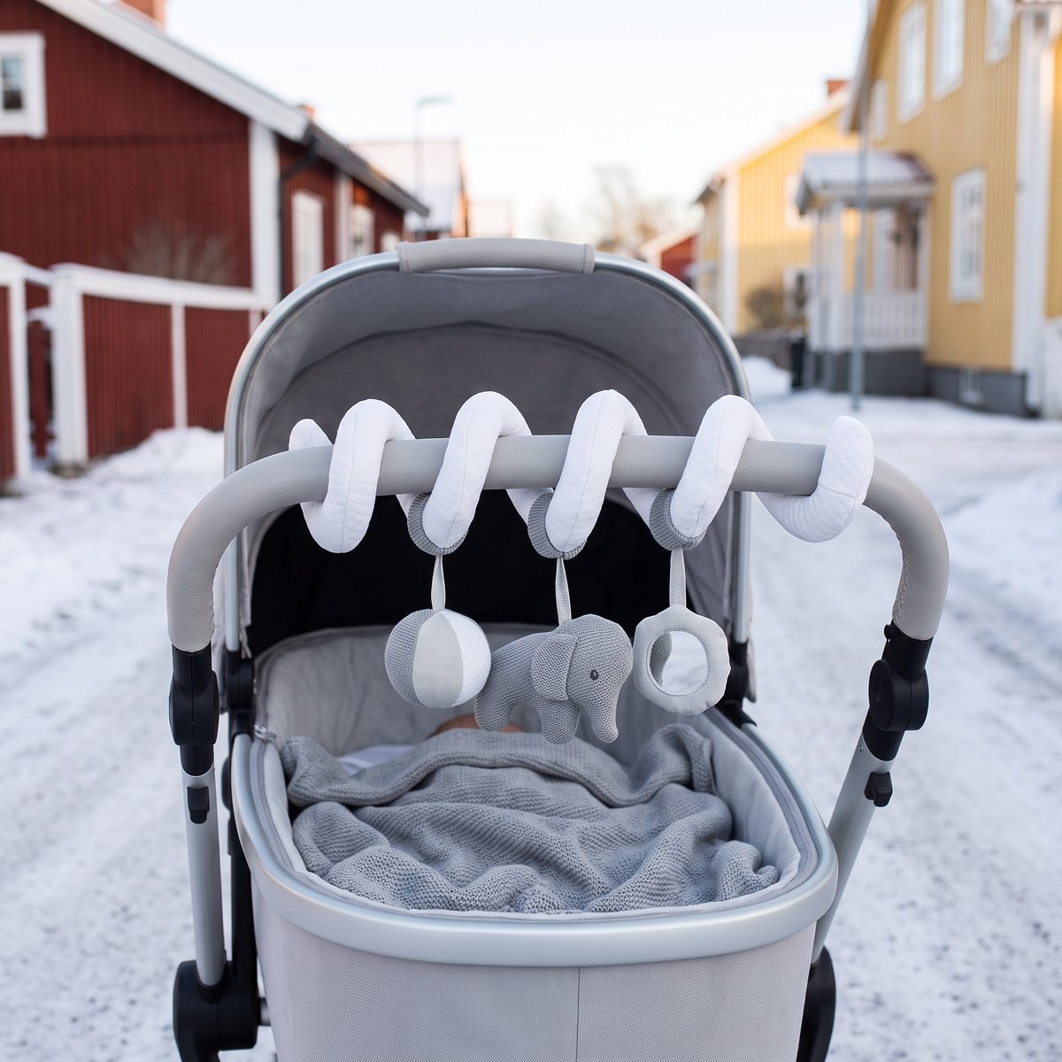 Nattou Tembo aktivitetsspiral lindad runt bygeln på en ljusgrå barnvagn på en snöig gata med röda och gula trähus