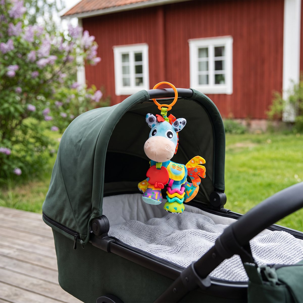 PlayGro Clip Clop hängd med sin krok i suffletten på en grön barnvagn utanför en röd stuga med syrener