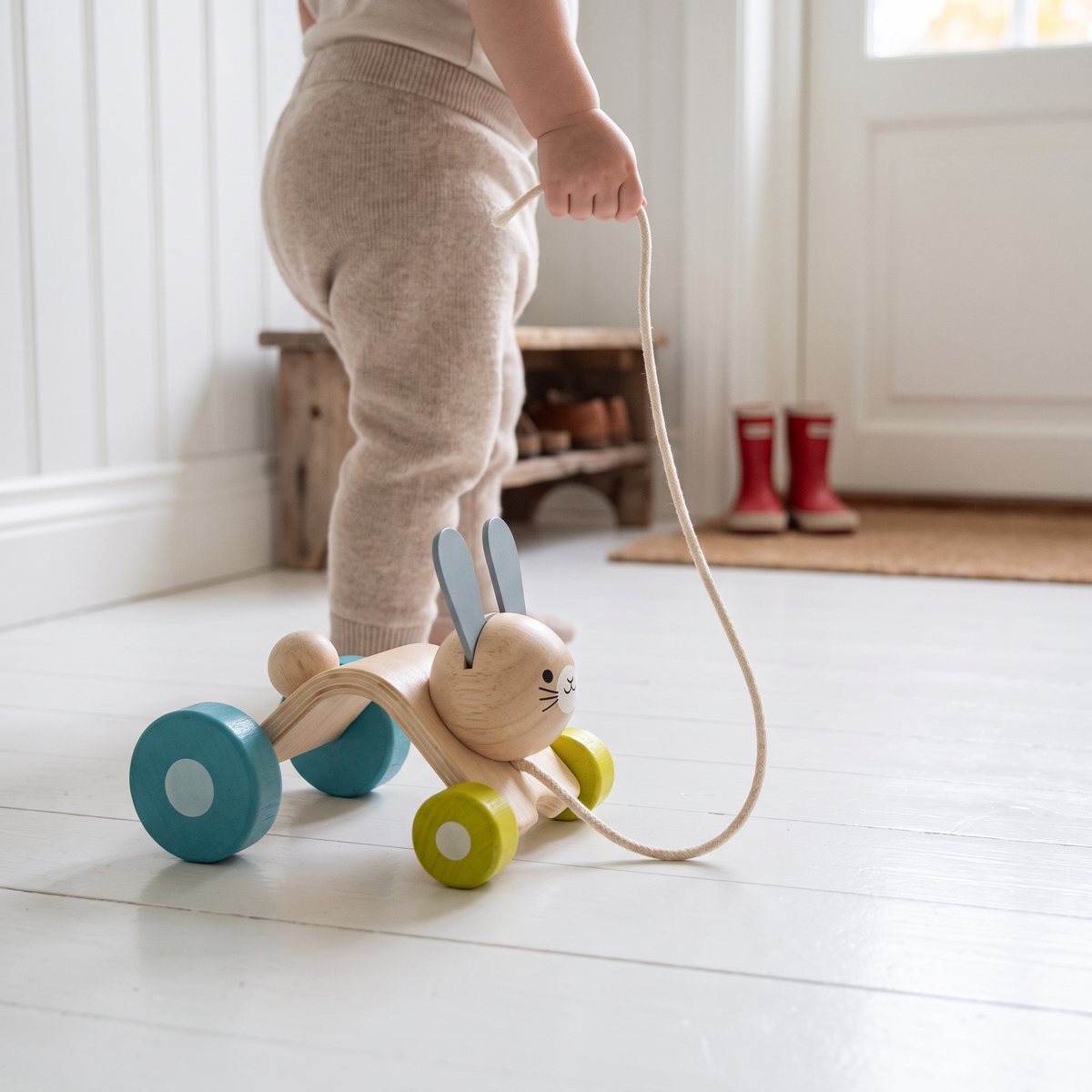 PlanToys Hopping Rabbit i trä med blå och gröna hjul som dras i snöret av ett litet barn i en ljus svensk hall