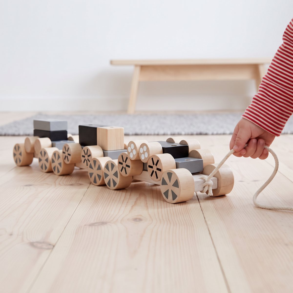 PlanToys Stacking Wheels, tre trävagnar med mönstrade hjul ihopkopplade till ett tåg, som en barnhand drar i snöret över ett ljust furugolv i ett svenskt barnrum