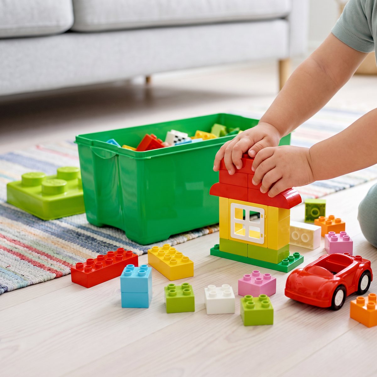 Barnhänder trycker fast DUPLO-klossar på ett litet hus med fönster bredvid den gröna förvaringslådan på ett svenskt vardagsrumsgolv