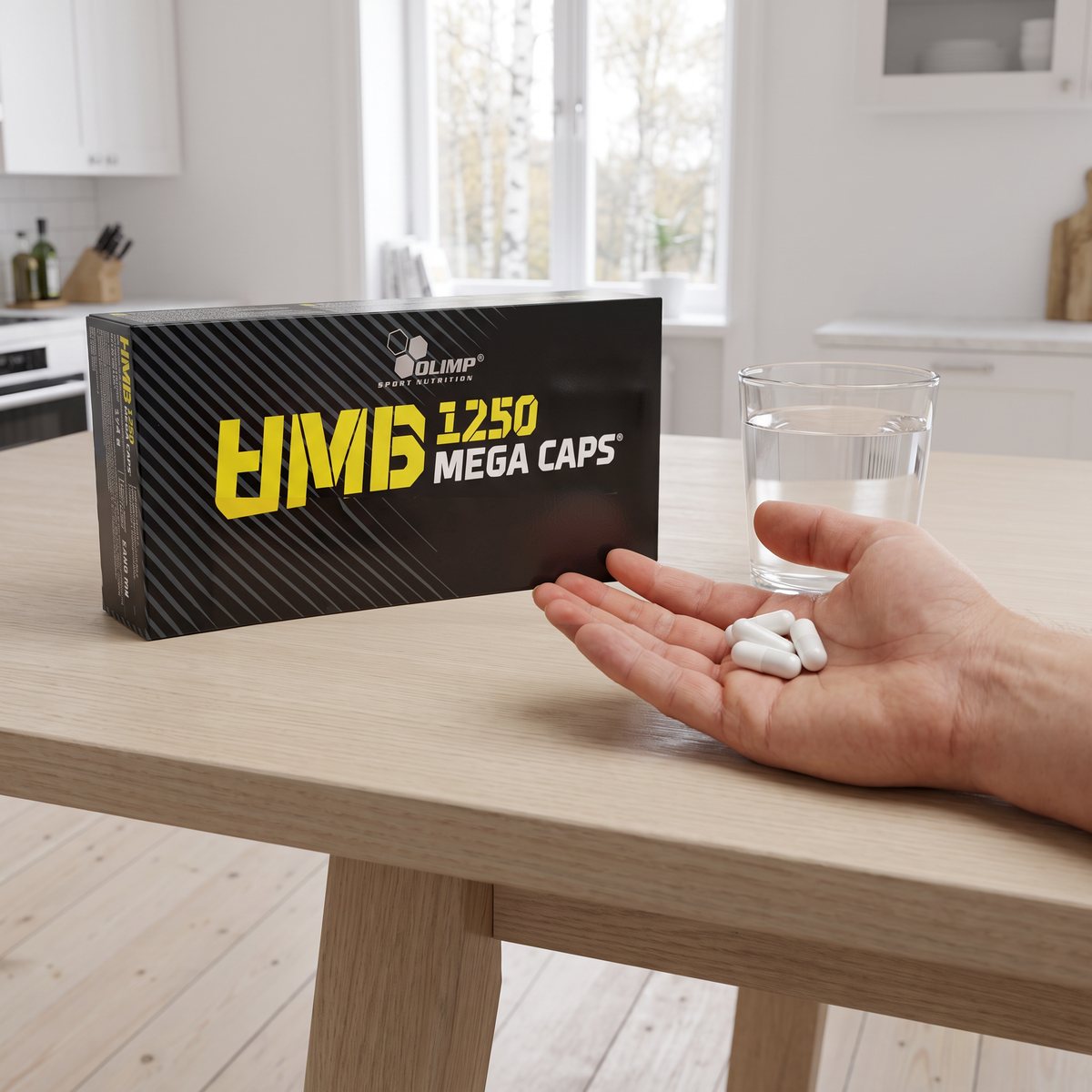 Olimp HMB Mega Caps-förpackning på ett köksbord i ljus ek med ett glas vatten och en hand som håller några kapslar, björkar utanför fönstret