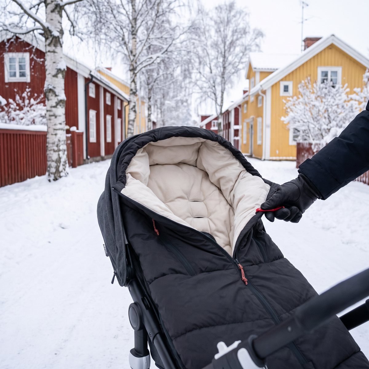 Svart Voksi Adventure-åkpåse med ljust foder i en sittvagn på en snöig gata med röda och gula trähus