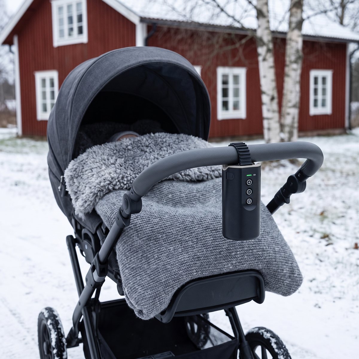 Svart Sleepytroll Baby Rocker Gen2 fastspänd på handtaget till en barnvagn som står ute i snön framför ett rött trähus med björkar