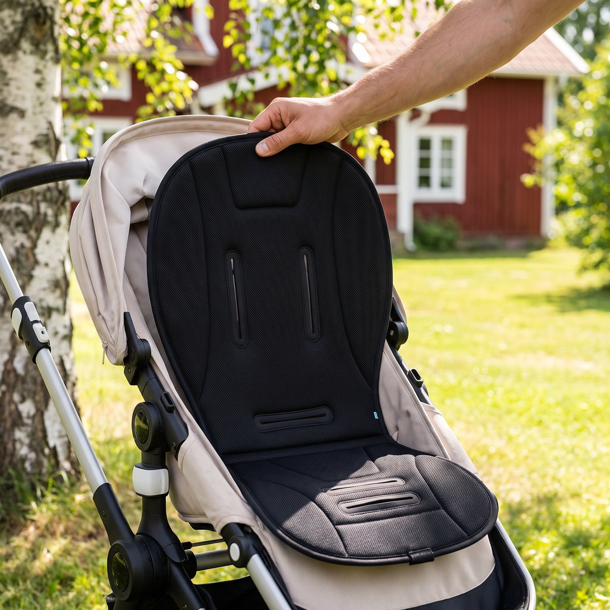 Svart sittdyna i 3D-mesh läggs i en beige barnvagn under en björk vid ett rött trähus en sommardag.