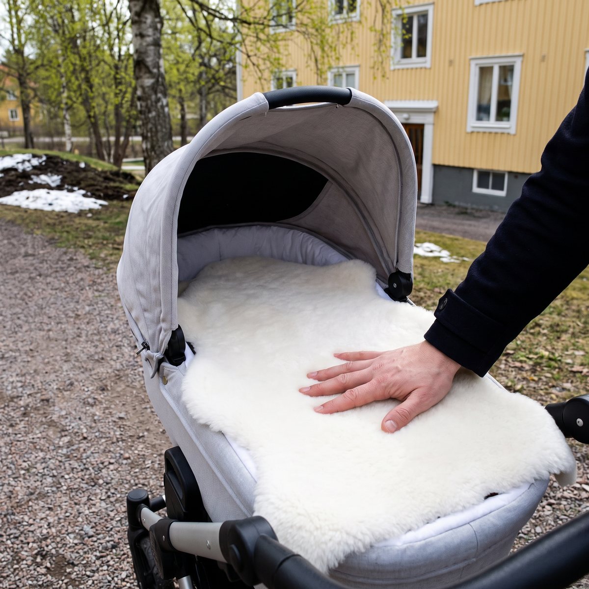 Vitt korthårigt lammskinn i liggdelen på en ljusgrå barnvagn, i en park med nyutslagna björkar en vårdag