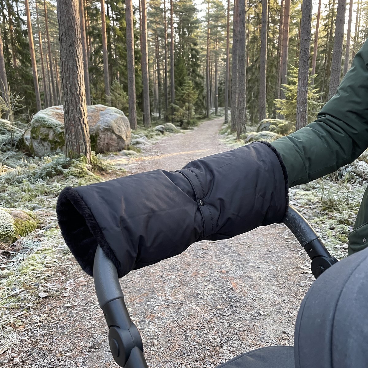 Svart fleecefodrad handmuff från Bozz över barnvagnens raka handtag, grusstig i en svensk tallskog med frost