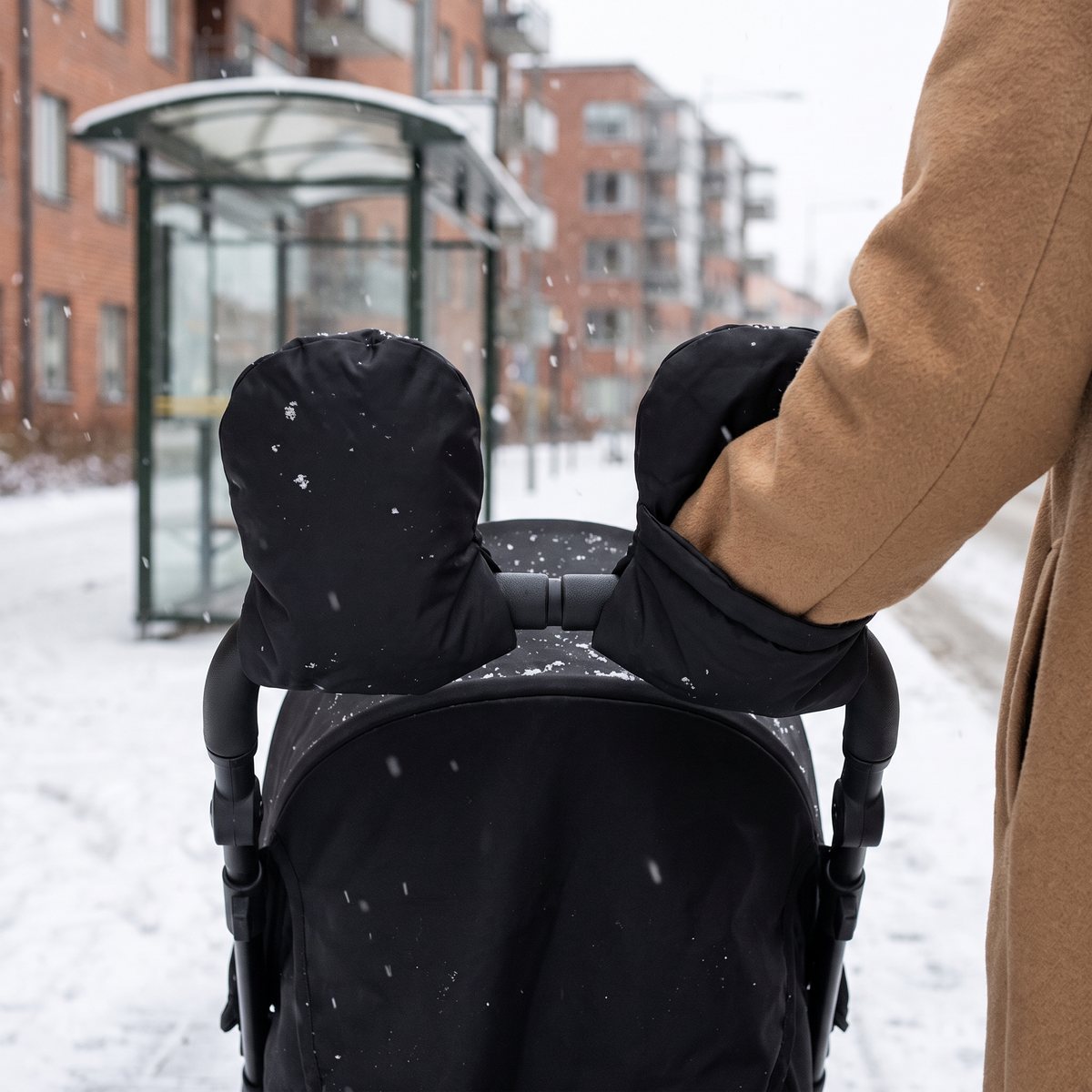 Små svarta barnvagnsvantar från Elodie på sittvagnens handtag vid en busskur i snöfall, svenska tegelhus i bakgrunden