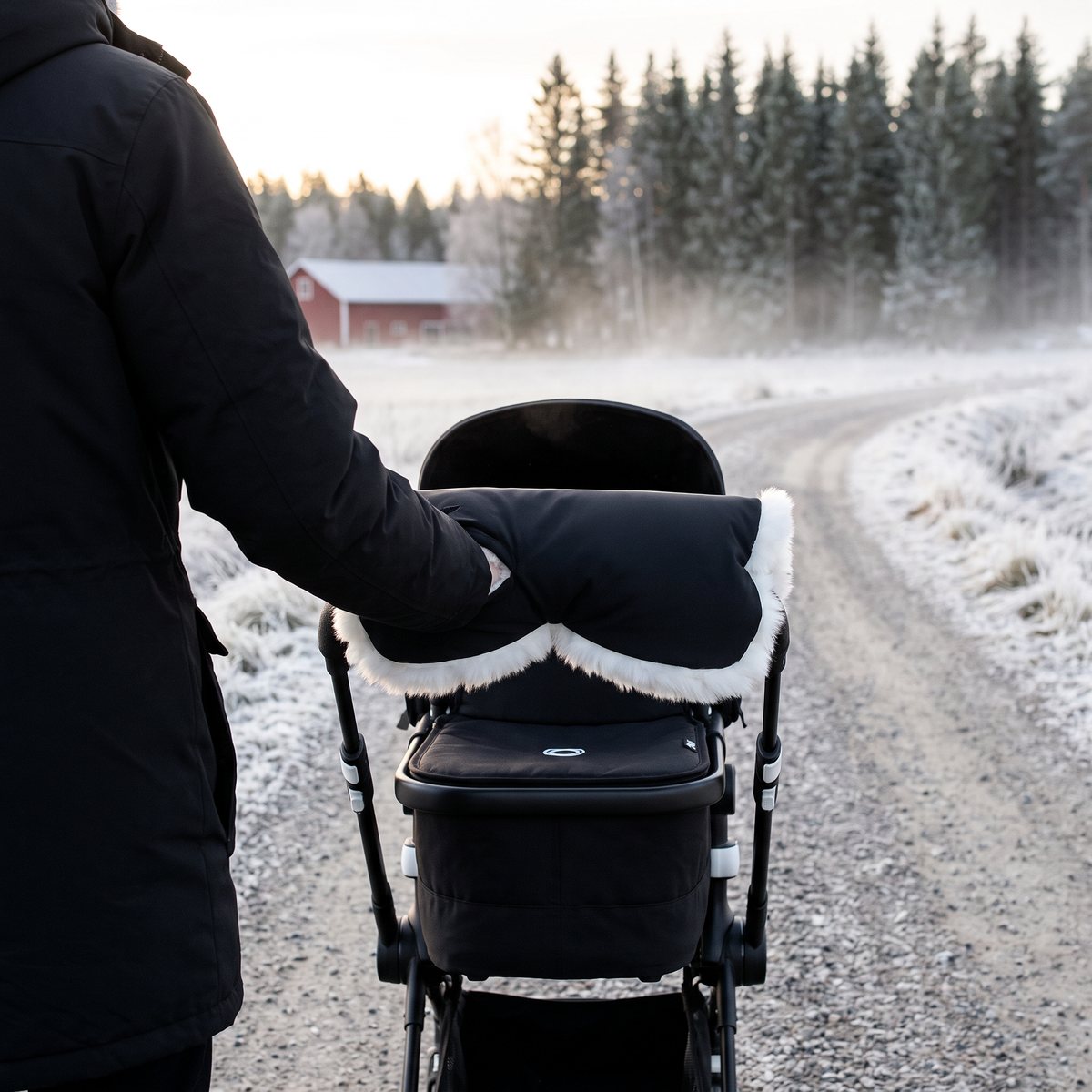 Svart handvärmare med vit lammskinnskant från Nordlys Thyra över barnvagnens handtag, frostig grusväg vid en åker med röd lada och granskog