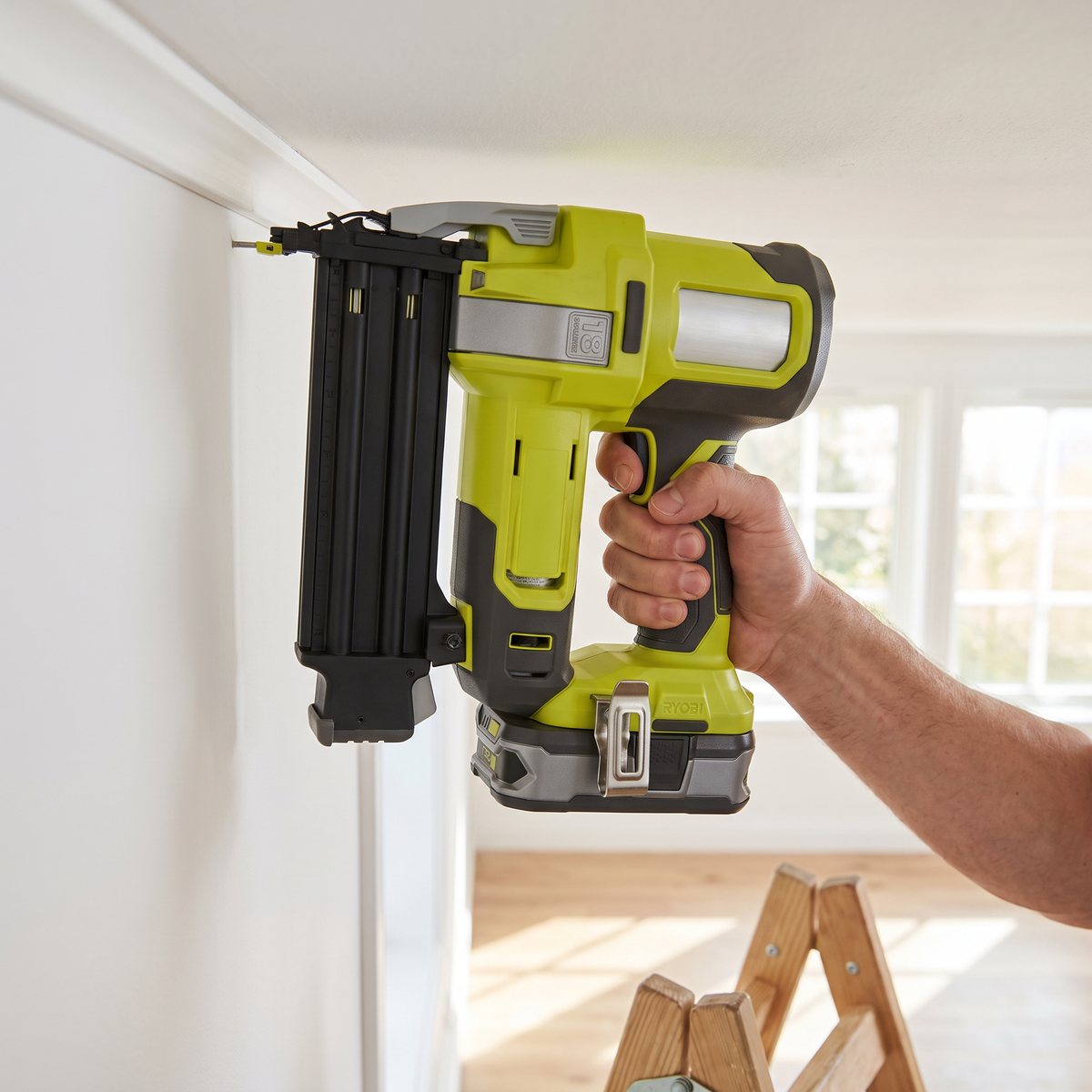 Limegrön batteridriven dyckertpistol som fäster en vit taklist i ett ljust svenskt vardagsrum, med en trappstege nedanför