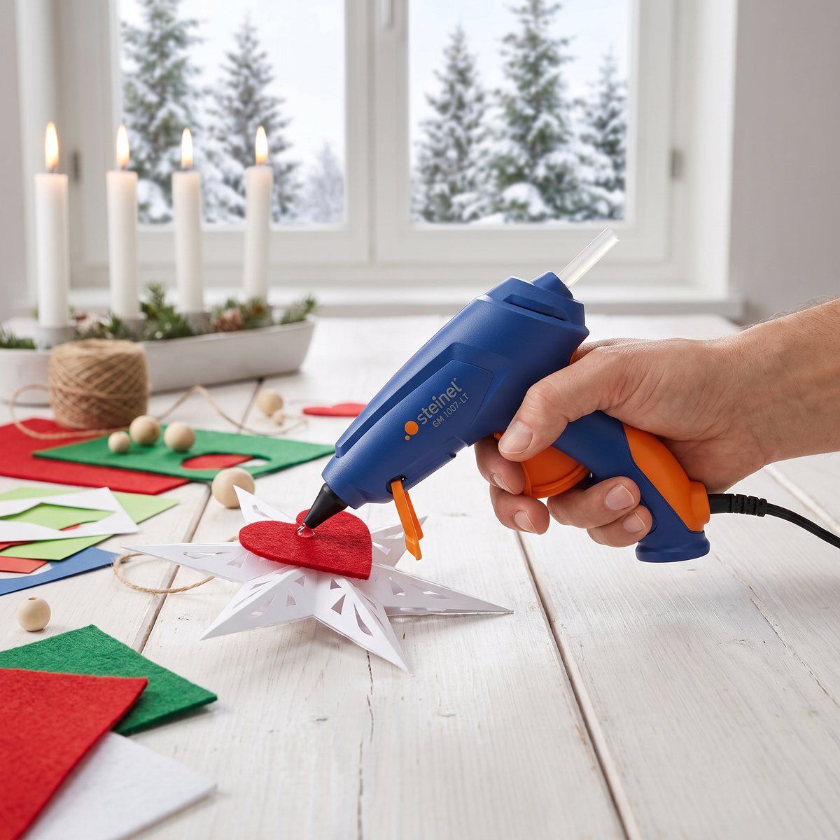 Blå och orange Steinel GlueMatic 1007LT som limmar ett filthjärta på en pappersstjärna vid ett adventspyssel med snöig gran utanför fönstret