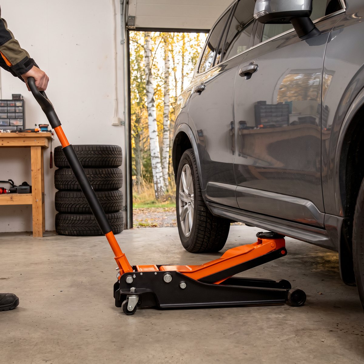 Orange och svart Bahco BH13000 garagedomkraft under tröskeln på en grå SUV i ett svenskt garage med betonggolv, vinterdäck och björkar utanför porten