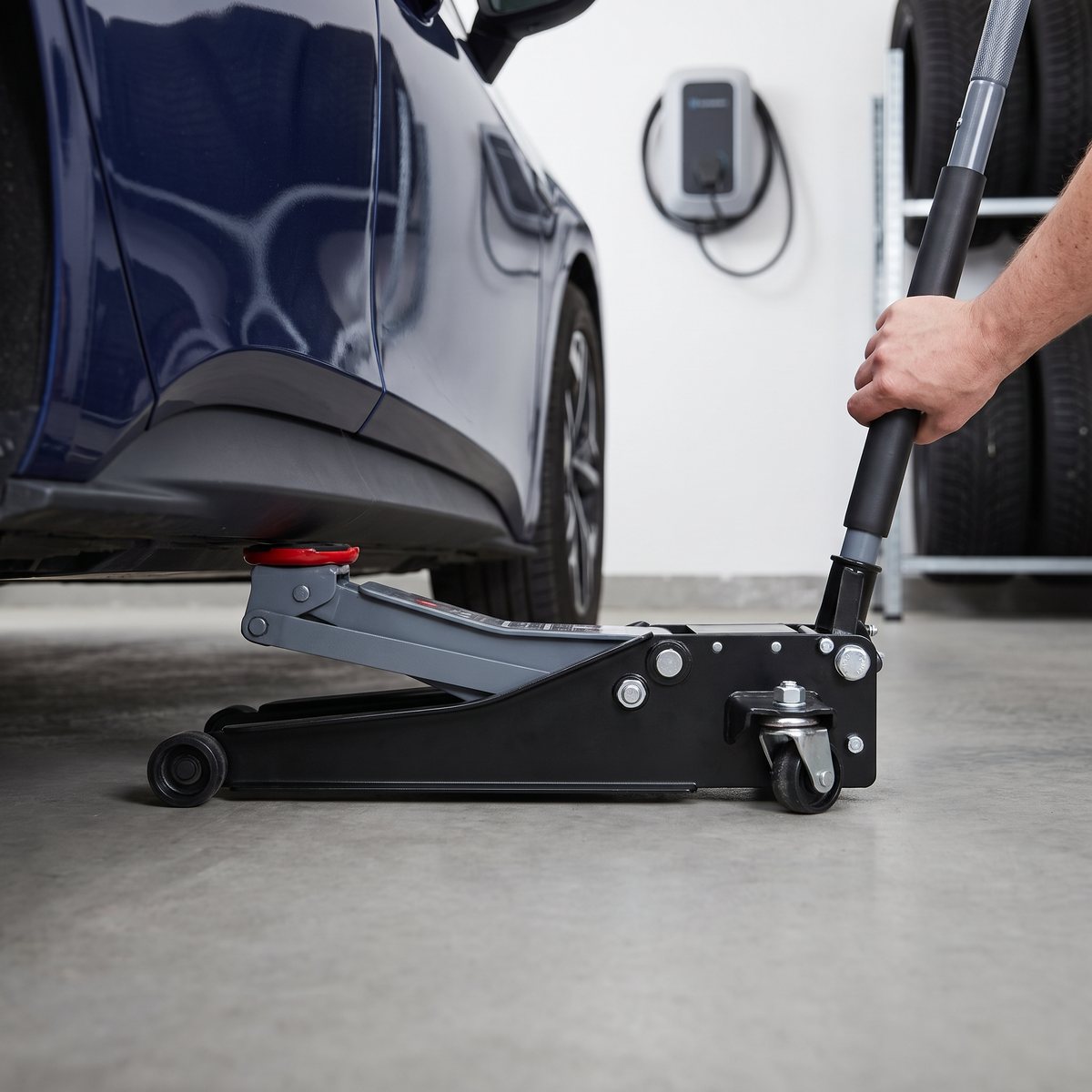 Svart lågprofil PELA 502993 garagedomkraft som rullas in under sidokjolen på en mörkblå elbil i ett svenskt garage med laddbox
