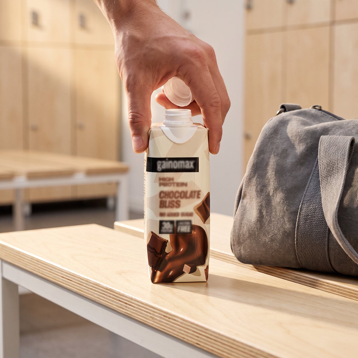 Gainomax High Protein Drink Chocolate i kartong på en träbänk i ett ljust omklädningsrum, bredvid en grå träningsväska