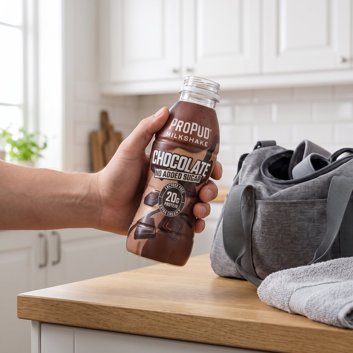 ProPud Protein Milkshake Chocolate i handen vid en köksbänk i ett ljust svenskt kök, bredvid en grå träningsväska och en handduk