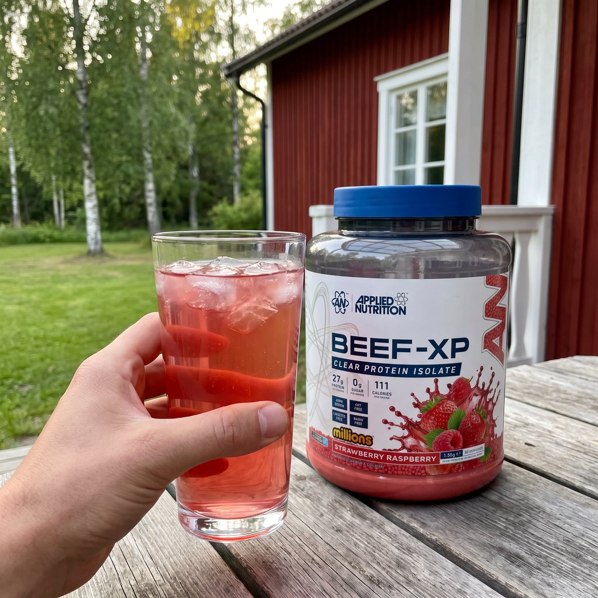 Burk med Applied Nutrition Beef XP Clear Protein Isolate bredvid ett glas klar röd dryck med is på verandan till en röd svensk sommarstuga