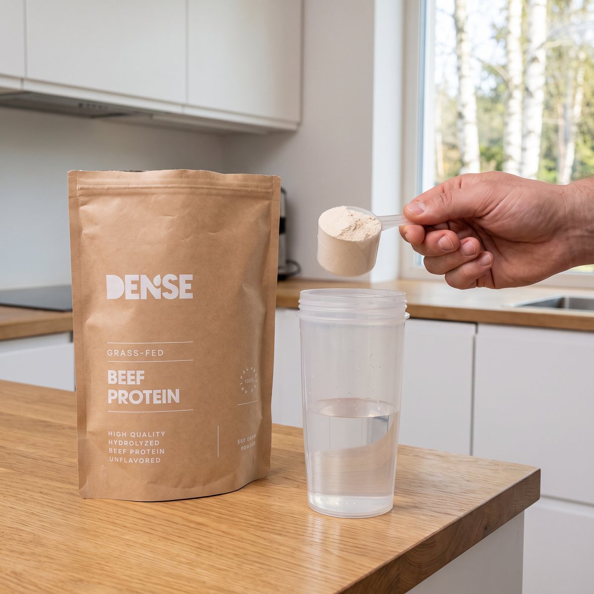Dense Biffprotein Naturell i kraftpapperspåse bredvid en shaker där en skopa pulver hälls i, i ett ljust svenskt kök med björkar utanför fönstret