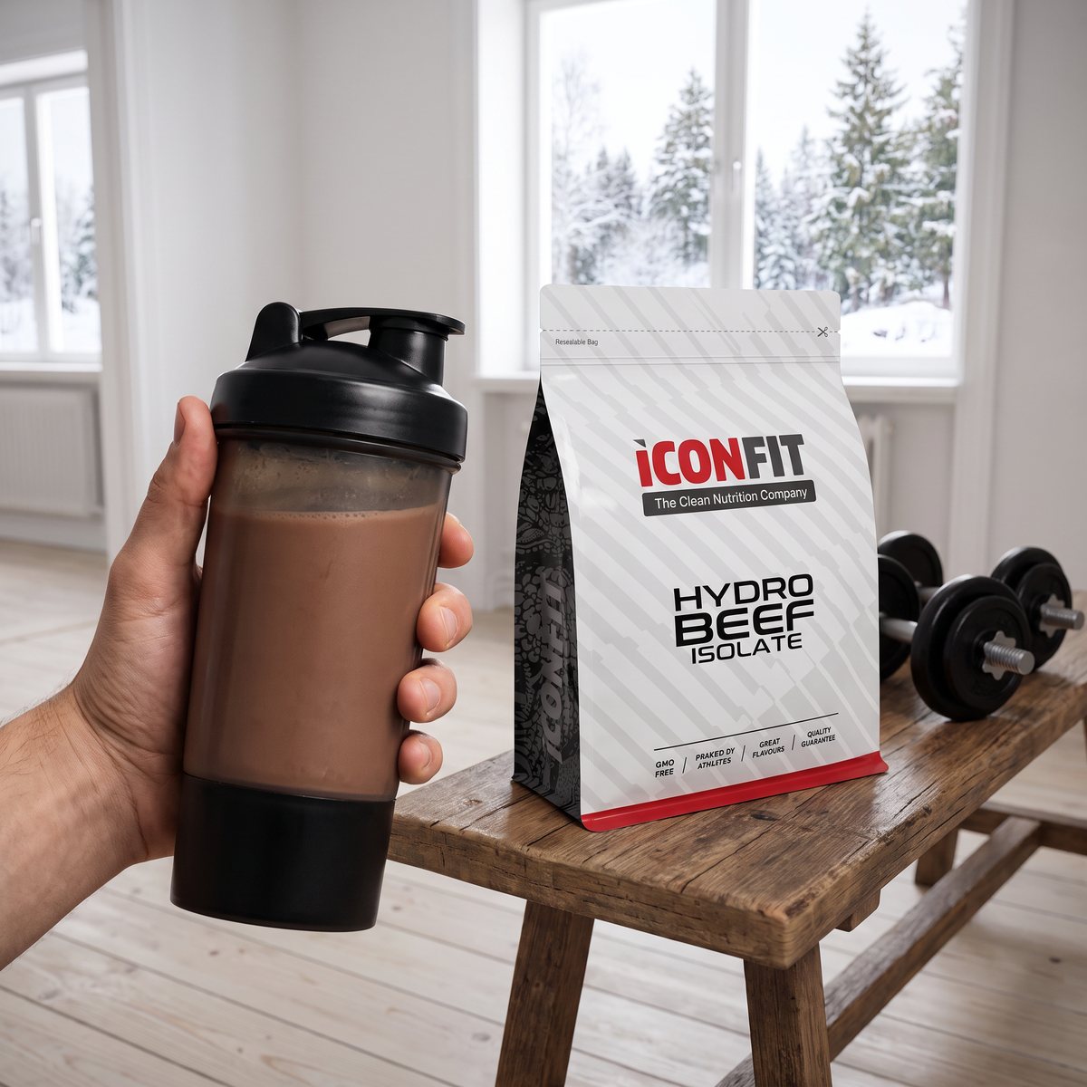 ICONFIT HydroBEEF Isolate-påsen på en träbänk bredvid hantlar och en shaker med chokladshake, i en ljus svensk lägenhet med snötäckta granar utanför