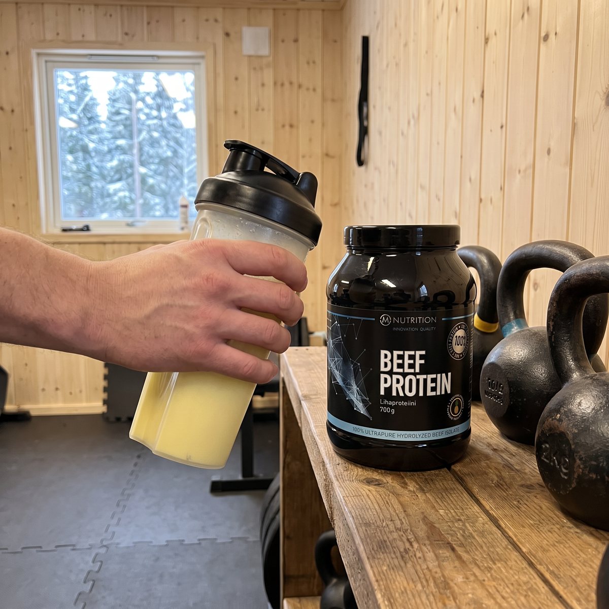 Svart burk med M-Nutrition Beef Protein på en trähylla bredvid kettlebells i ett svenskt garagegym, en hand håller en shaker med gul dryck