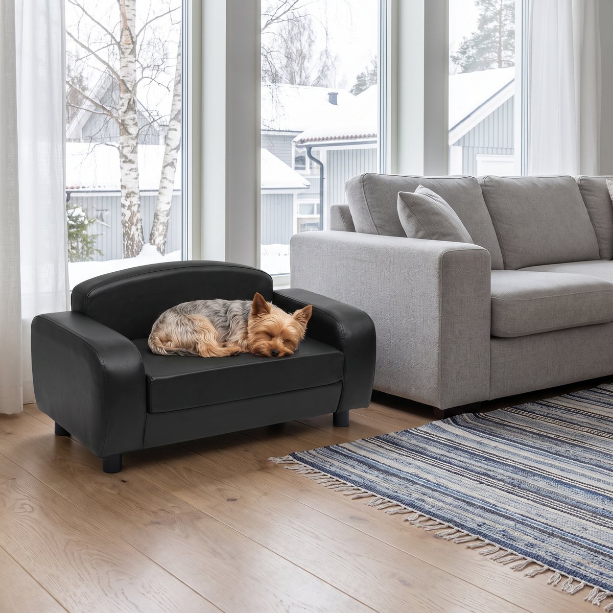 Svart hundsoffa i konstläder med armstöd och korta ben bredvid en grå soffa i ett vardagsrum, en yorkshireterrier ligger hoprullad på sitsen