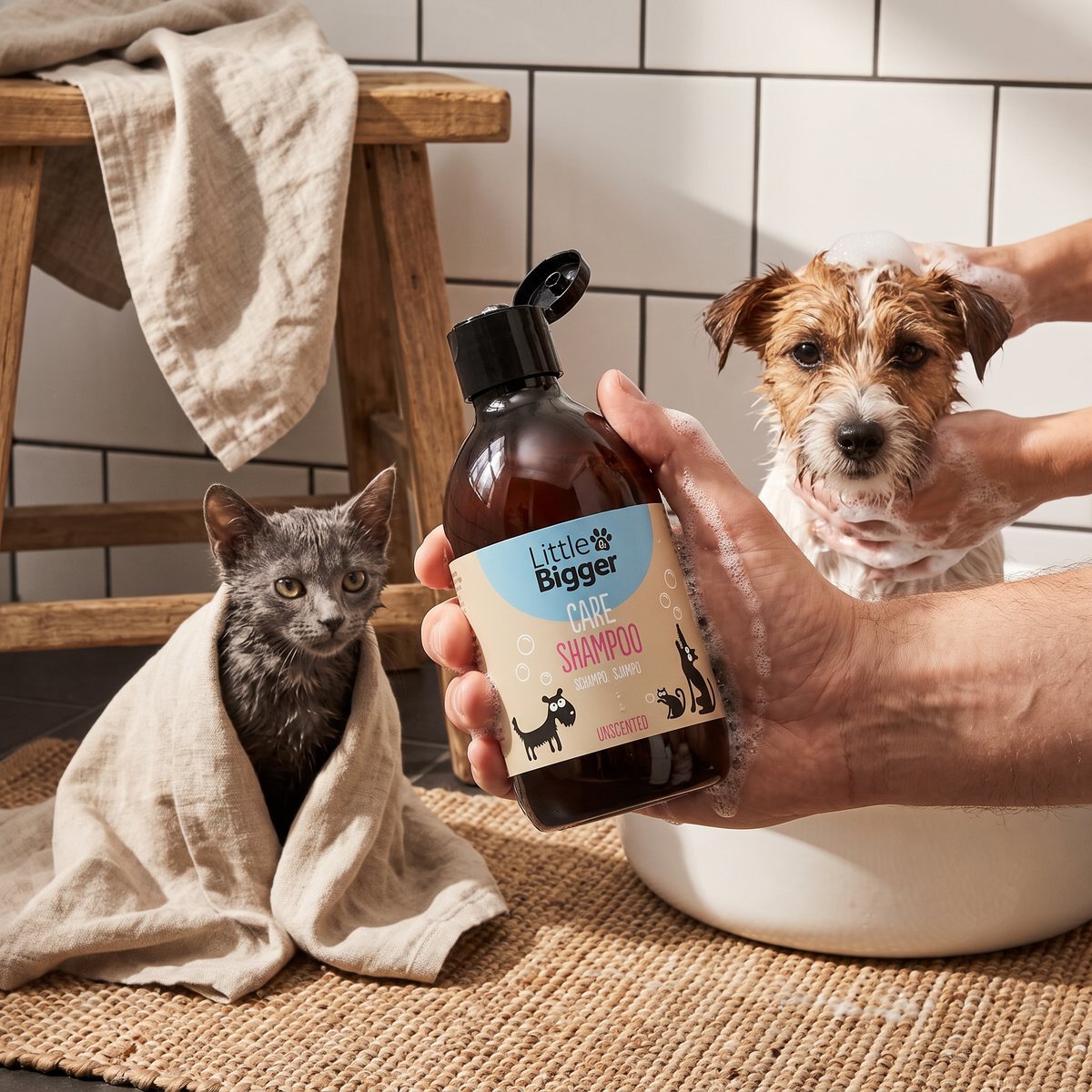 En katt inlindad i en linnehandduk och en terrier som tvålas in i en balja, med en hand som håller Little&Bigger Care-flaskan