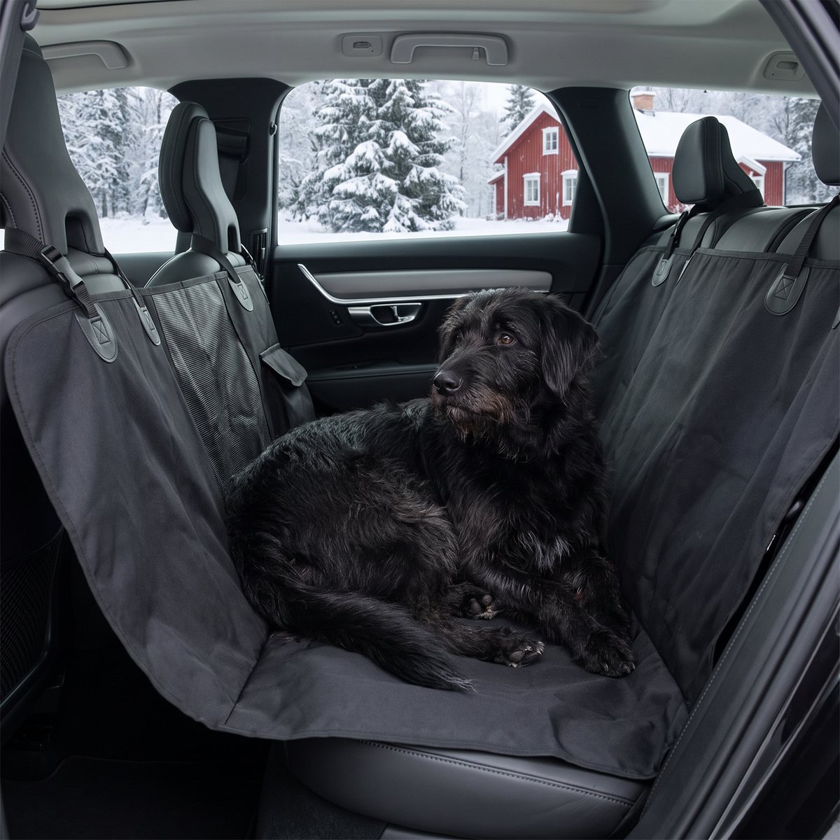 Svart baksätesskydd i oxfordtyg som hammock i en familjebil med en svart hund, snötäckta granar och rött hus utanför
