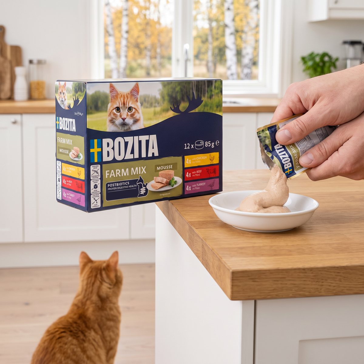Bozita Mousse Farm Mix-förpackning på en träbänk i ett ljust svenskt kök medan en hand klämmer ljus mousse ur en påse ner i en skål, med en rödrandig katt som väntar