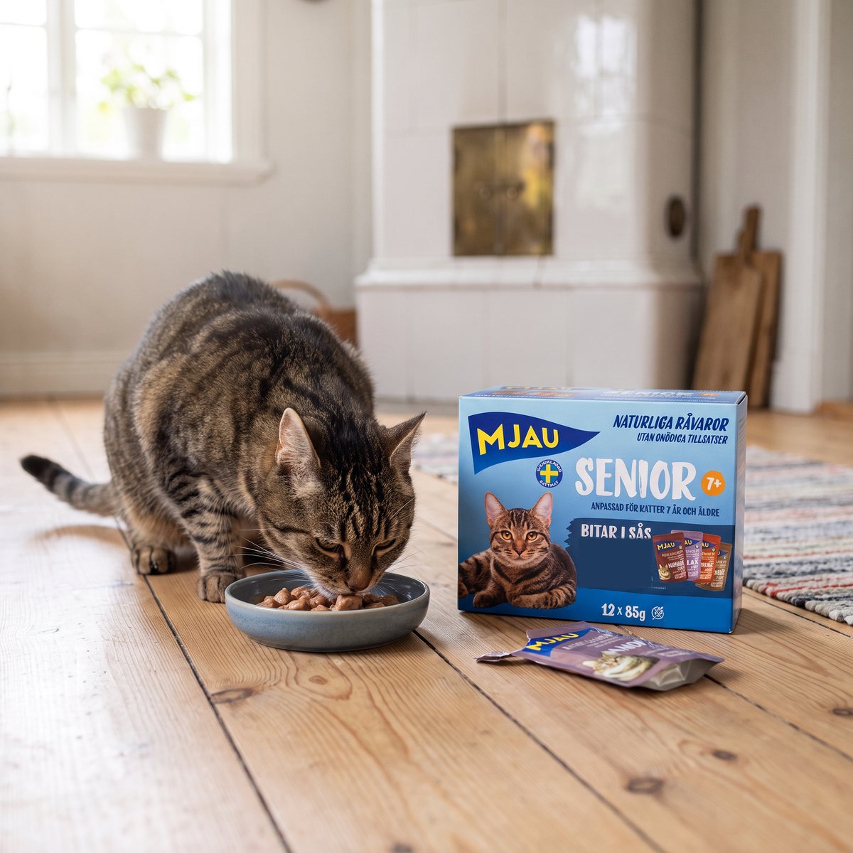 En äldre tigrerad katt äter bitar i sås ur en skål bredvid en Mjau Multibox Senior-låda på ett trägolv i ett svenskt lantkök med kakelugn
