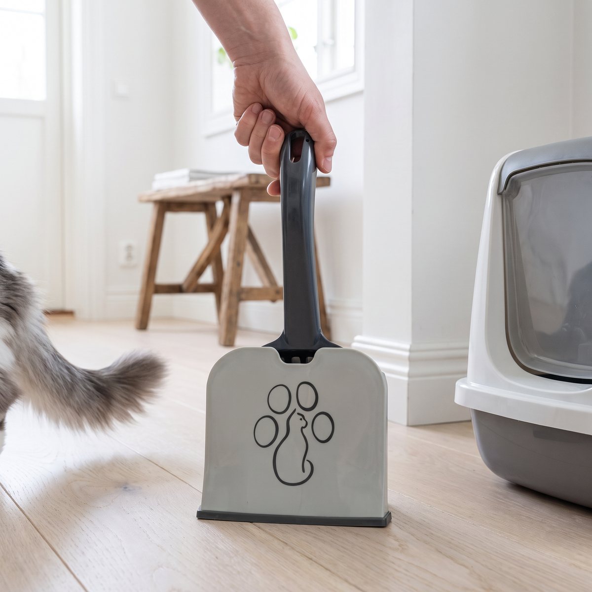 Kattsandsspade med ställ i grått med tassmotiv, hållen i handtaget i en svensk hall