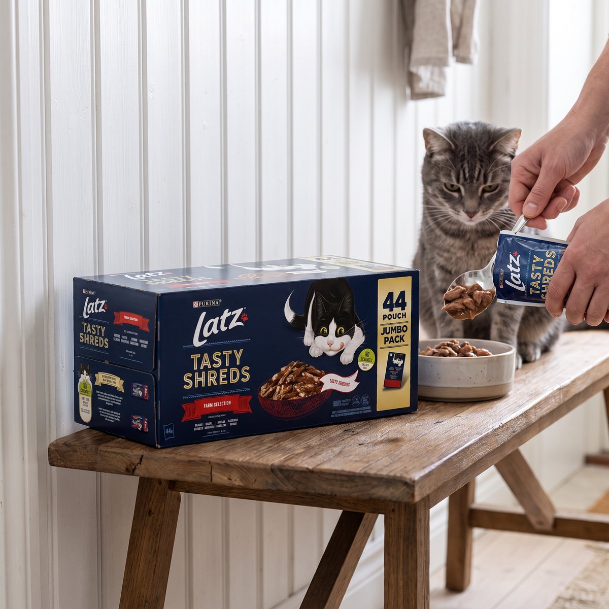 Kartong med Latz Tasty Shreds Farm Selection på en träbänk i en svensk hall, en hand häller strimlor i en kattskål