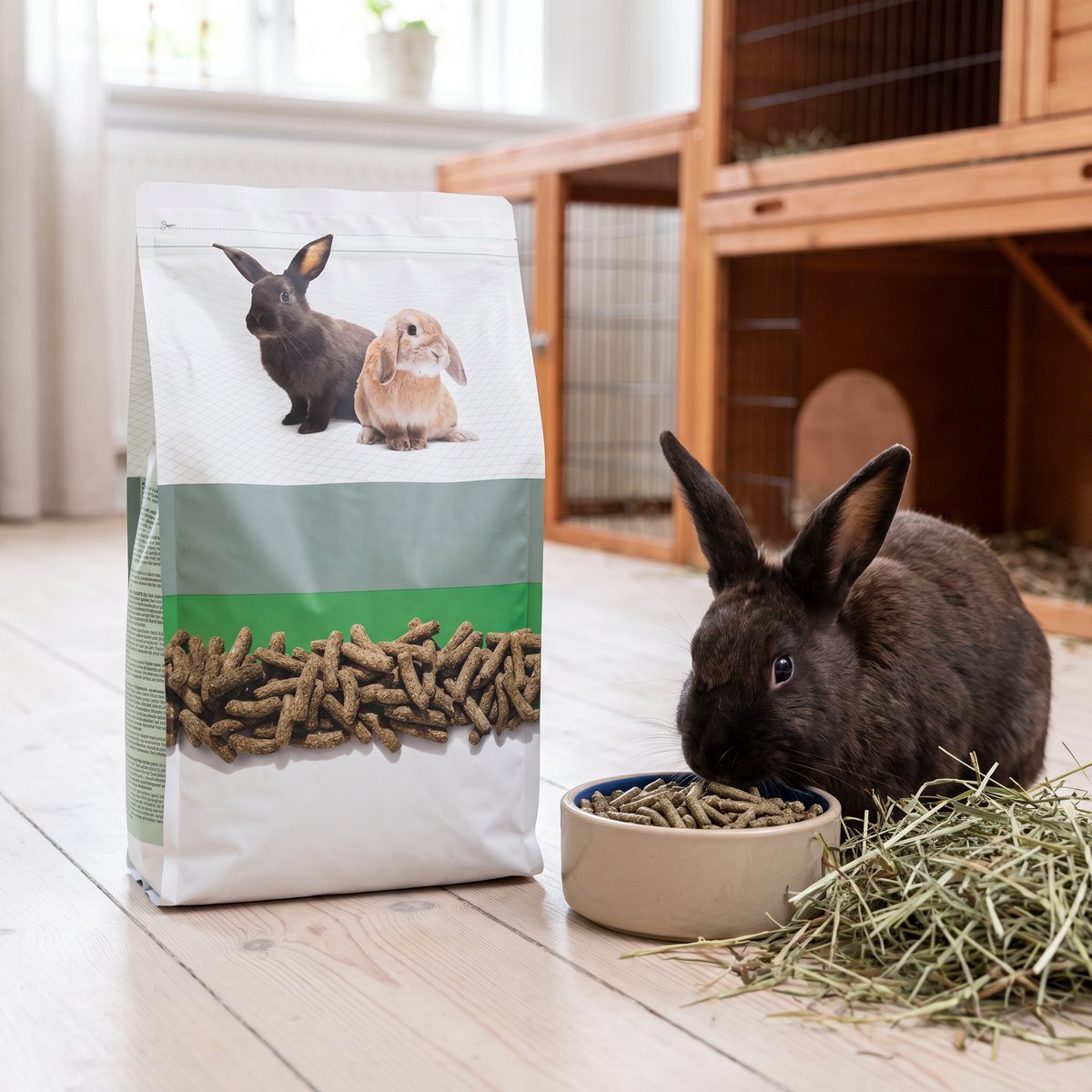 Versele-Laga Complete Cuni Adult-påse på ljust trägolv i ett svenskt hem, bredvid en mörk kanin som äter pellets ur en skål med hö intill