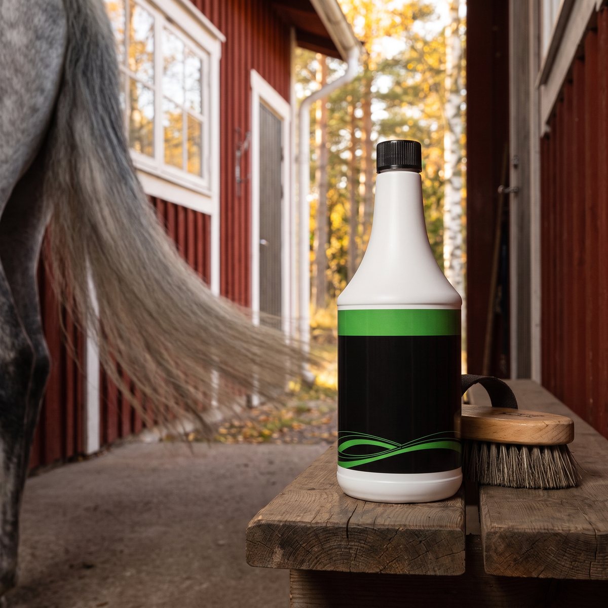 Flaska med Trikem Mane & Tail på en träbänk vid en grå hästs svans utanför ett rött stall