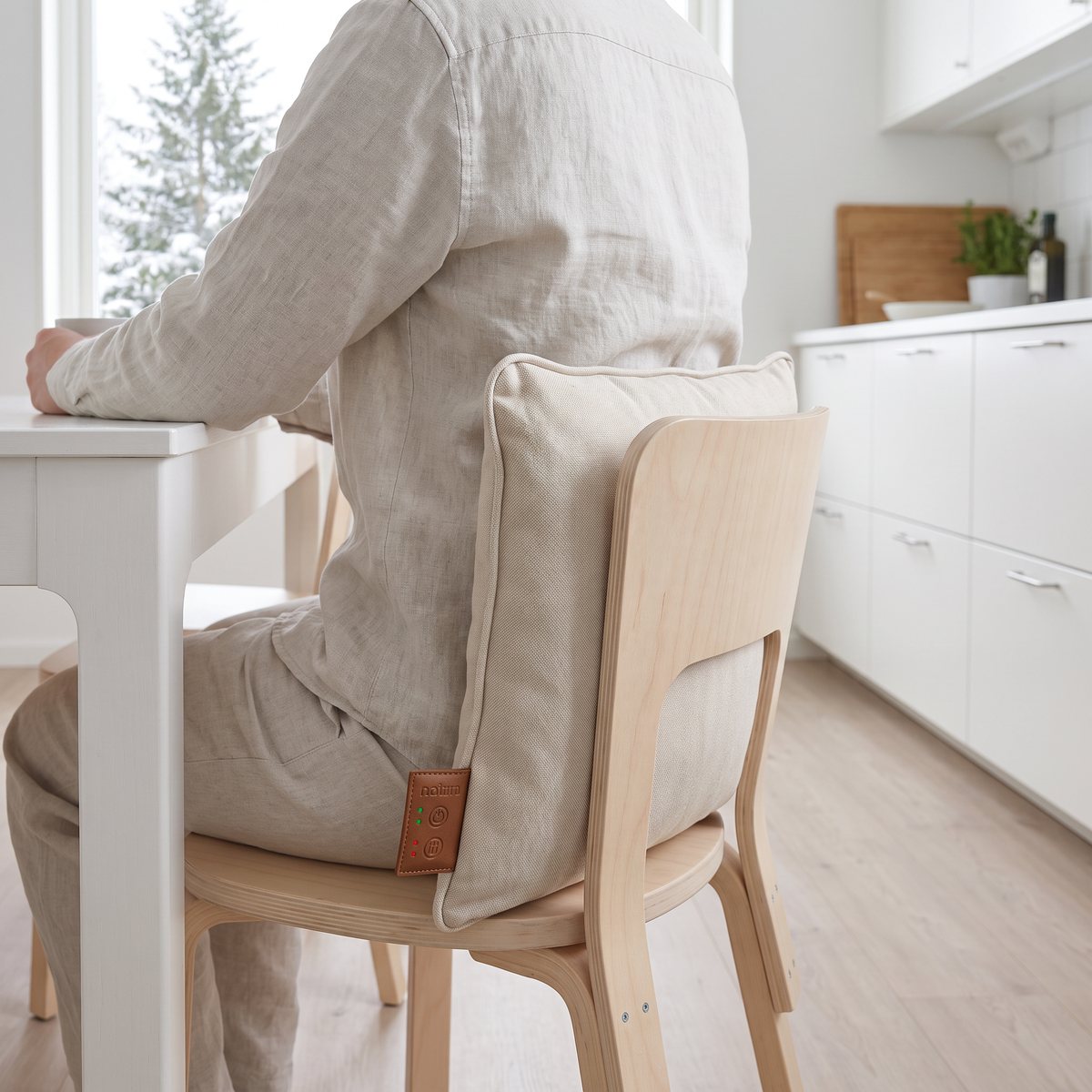 Beige massagekudde med värme mot ryggen på en björkstol vid ett vitt köksbord i ett svenskt hem